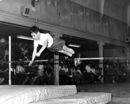 A young boy with his arms stretched out in front of him, is suspended in midair, almost clearing the bar of the high jump.  His sneakered feet are still on the other side of the bar.  He will land on two padded mats.